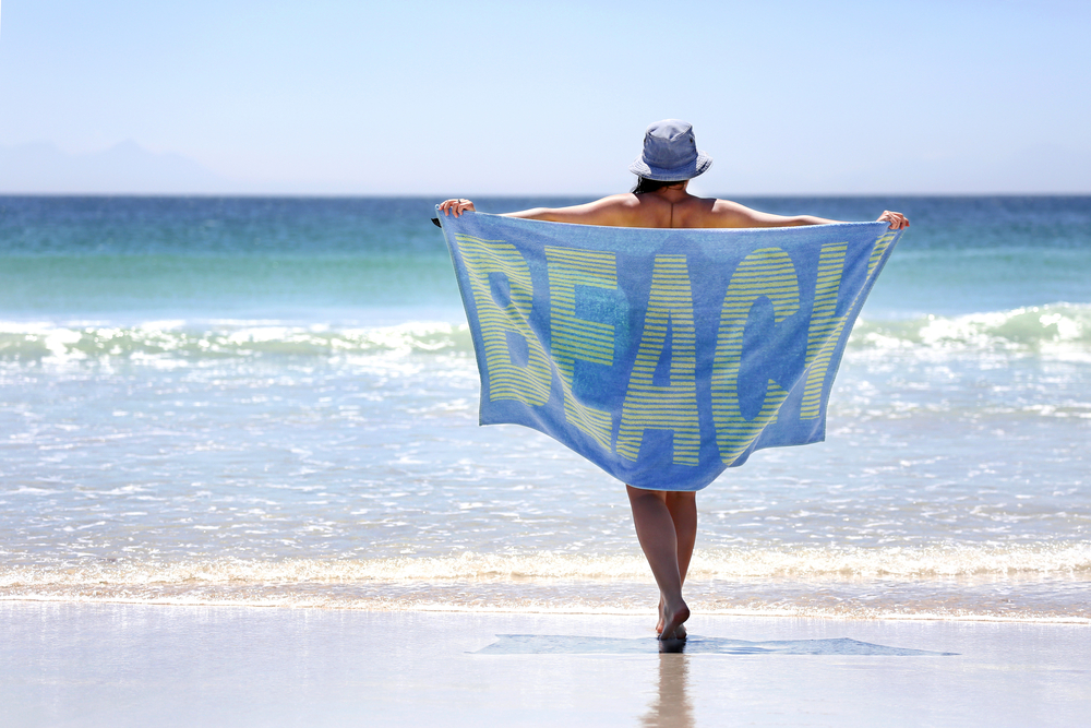 Woman walking with a towel around her on a pristine beach with the word BEACH spelled out on the towel.